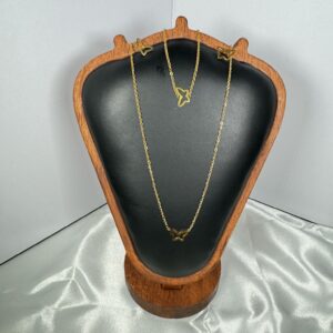 A gold-toned jewelry set featuring a delicate butterfly silhouette pendant necklace and matching stud earrings, elegantly displayed on a black leather and wood stand against a moody blue background.