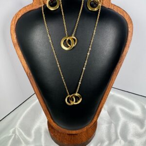 A gold-toned jewelry set featuring an interlocking double-circle pendant necklace and matching circle drop earrings, displayed elegantly on a dark leather and wood jewelry stand against a royal blue background.