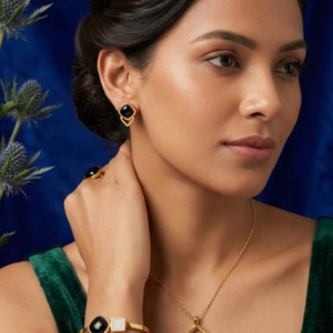 A woman with an elegant updo wearing the Onyx geometric gold pendant necklace, matching black onyx earrings, a crystal-accented bangle, and a coordinating ring, styled with a sophisticated dark green velvet dress against a deep blue background.
